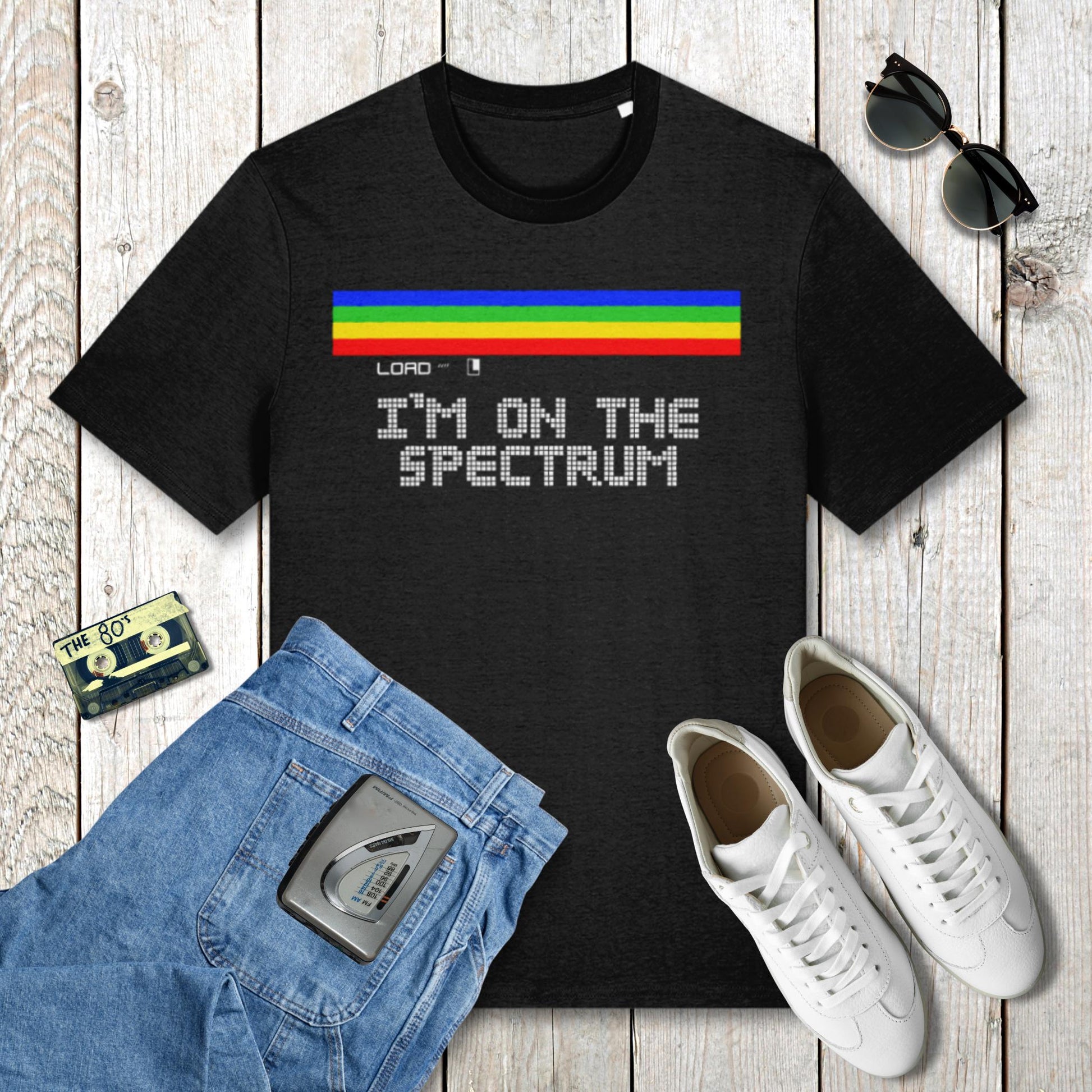 Black t-shirt with 'I'm on the spectrum' text and rainbow stripe, placed on a wooden surface with jeans, sneakers, and sunglasses.