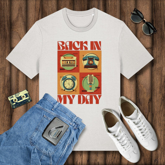 White t-shirt with 'Back in My Day' design, blue jeans, white sneakers, and sunglasses on a wooden surface.