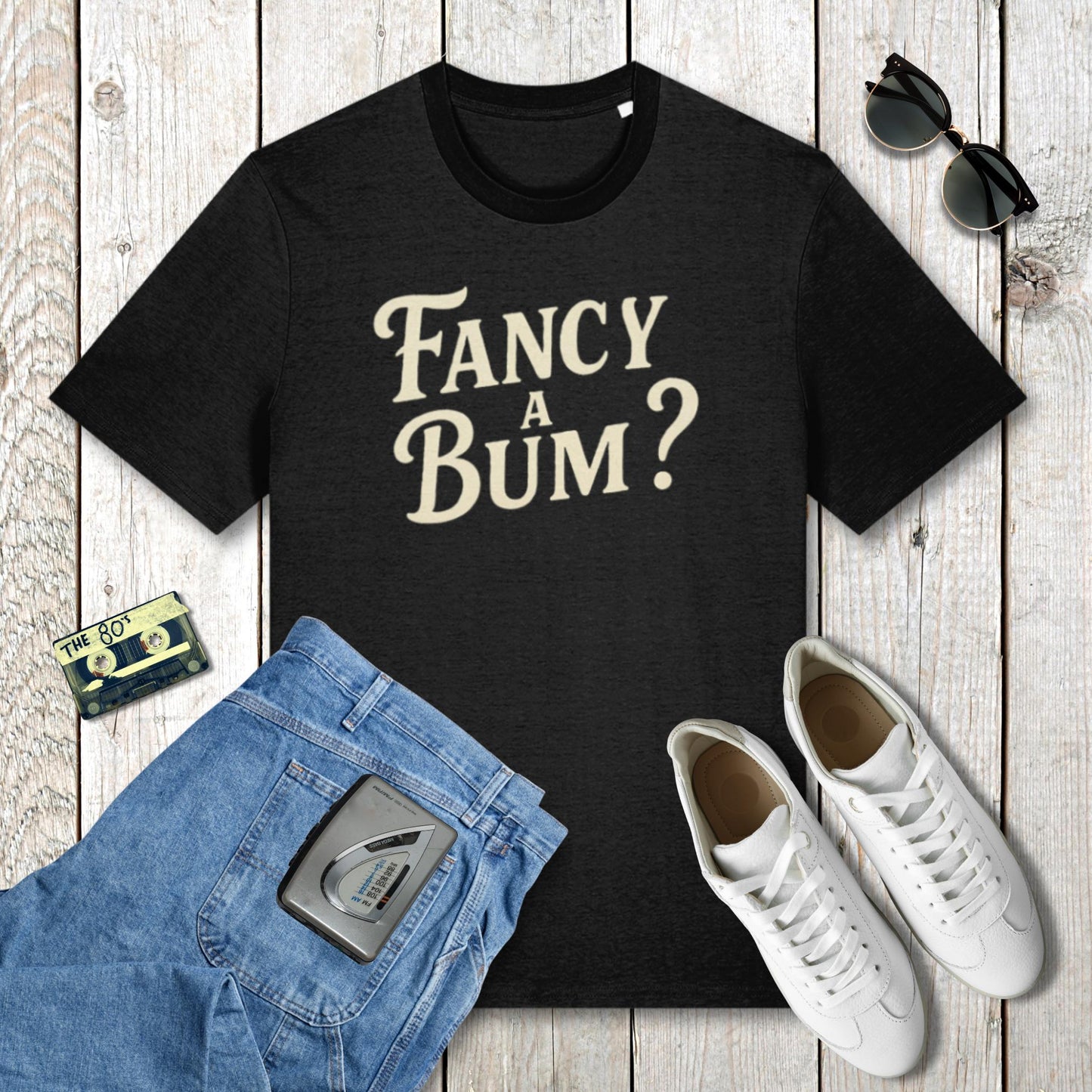 Black t-shirt with 'Fancy A Bum?' text, blue jeans, white sneakers, and sunglasses on a wooden surface.
