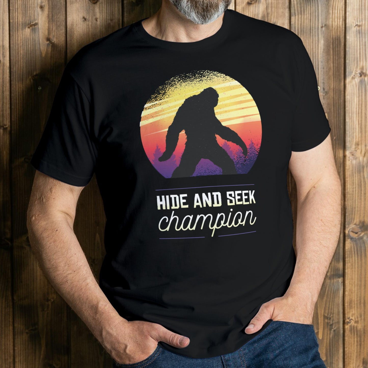man wearing a black t-shirt with a Sasquatch graphic and 'Hide and Seek Champion' text against a wooden background