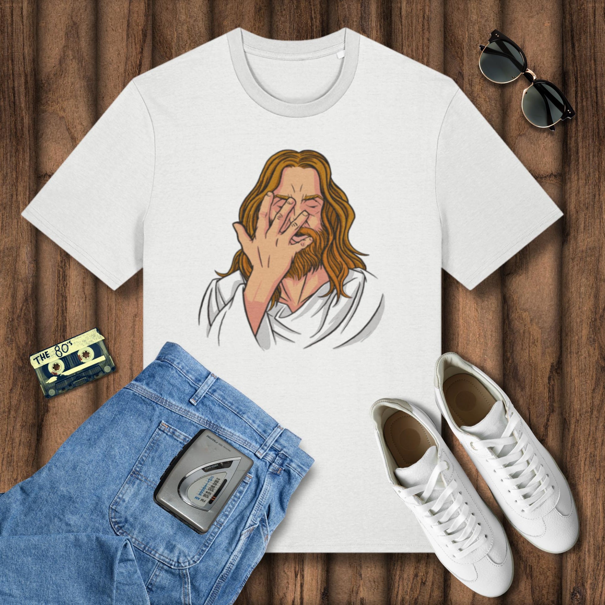 White t-shirt with a graphic design of a person covering their face, placed on a wooden surface with jeans, sneakers, and a cassette tape.