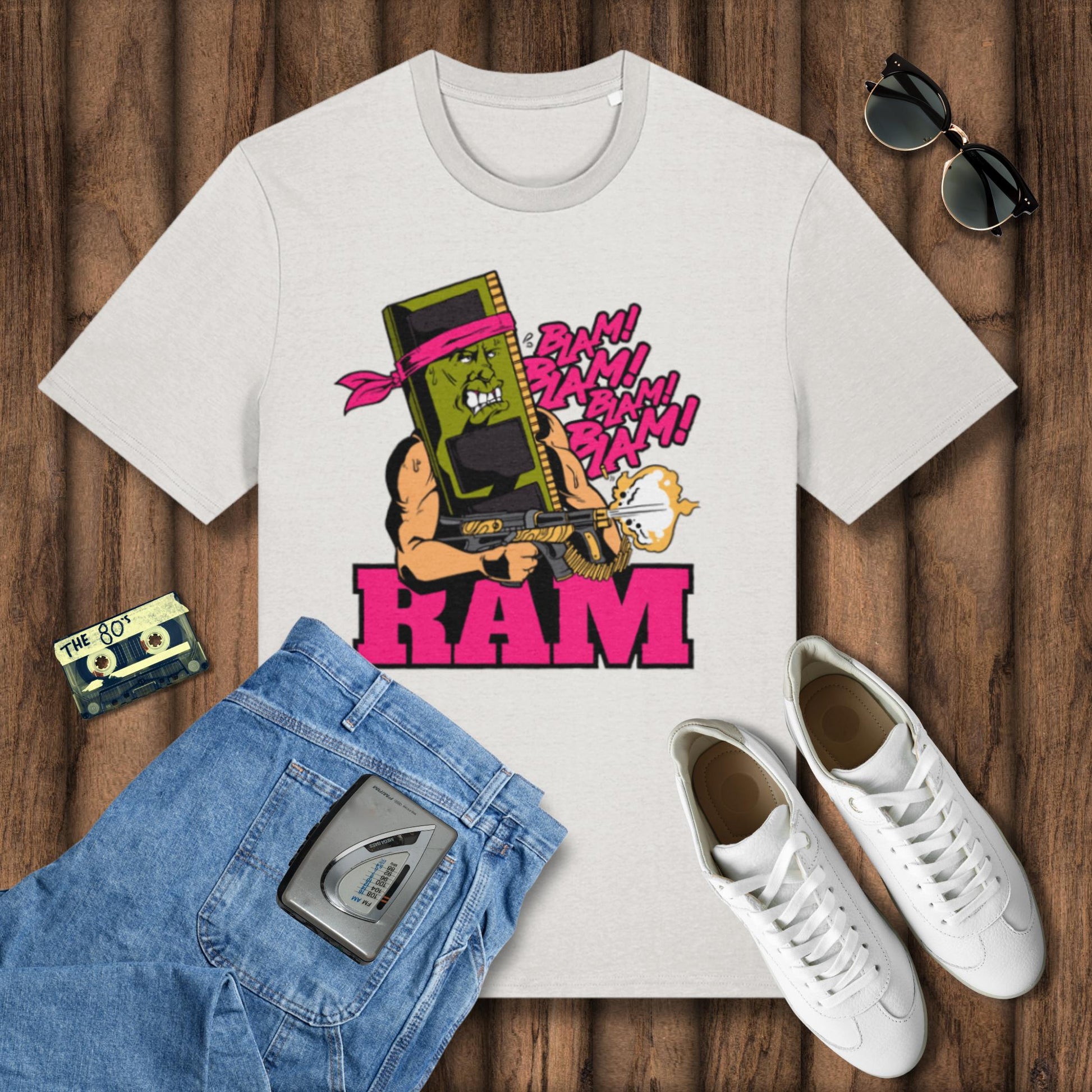 White t-shirt with graphic design of a character holding a boombox, surrounded by jeans, sneakers, and a cassette tape on a wooden surface.
