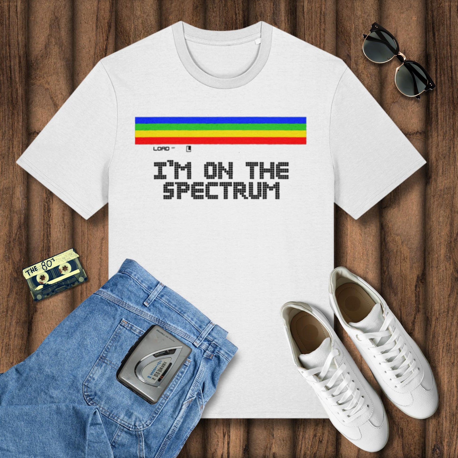 White t-shirt with 'I'm on the Spectrum' design, blue jeans, white sneakers, and sunglasses on a wooden surface.