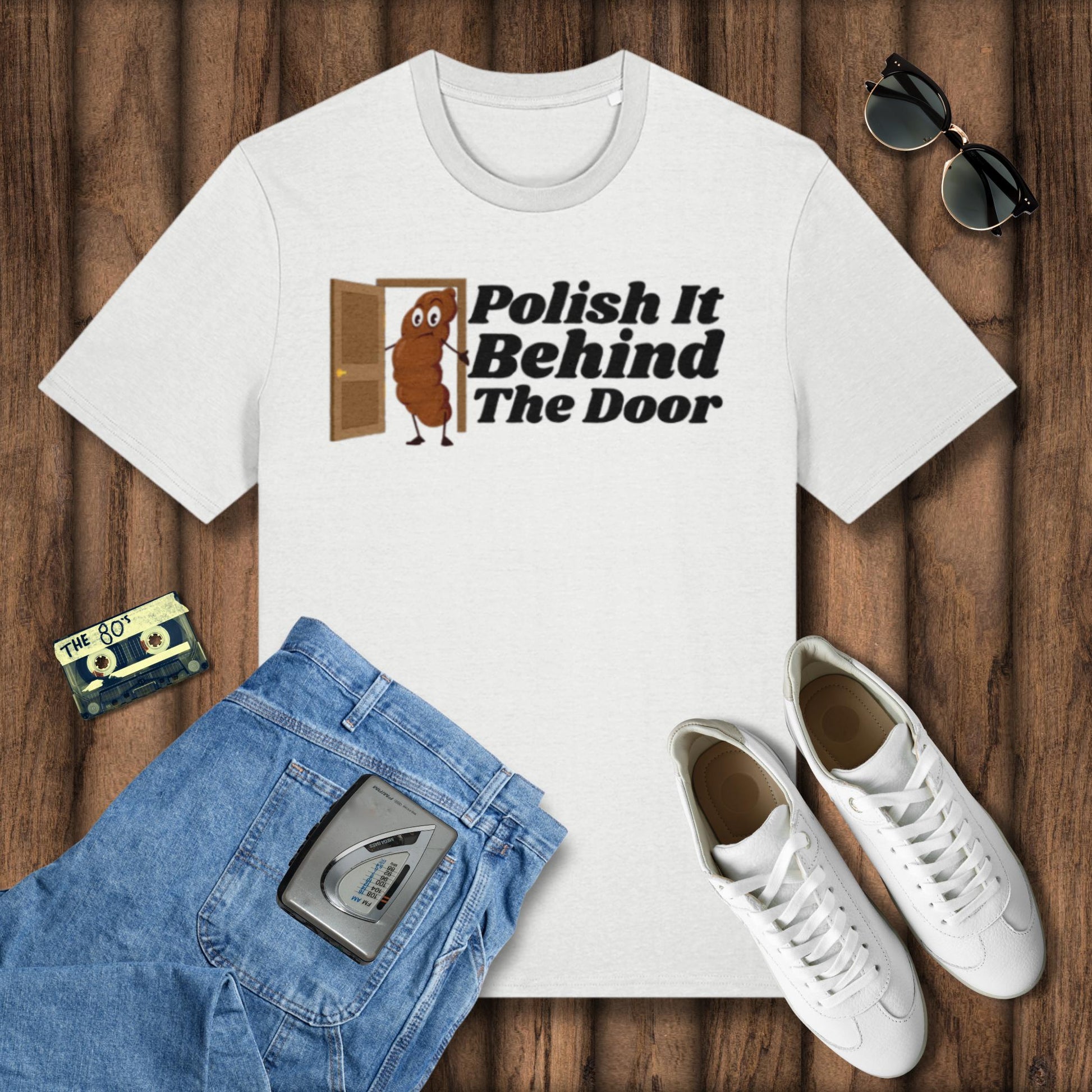White t-shirt with 'Polish It Behind The Door' graphic, blue jeans, white sneakers, and sunglasses on a wooden surface.
