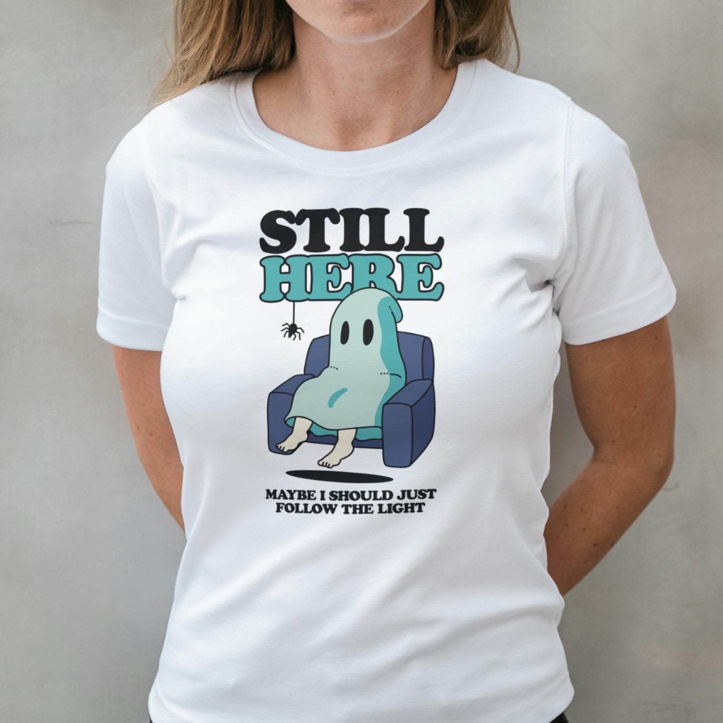 Person wearing a white t-shirt with a graphic of a ghost in a chair and text 'Still Here, Maybe I Should Just Follow the Light'.