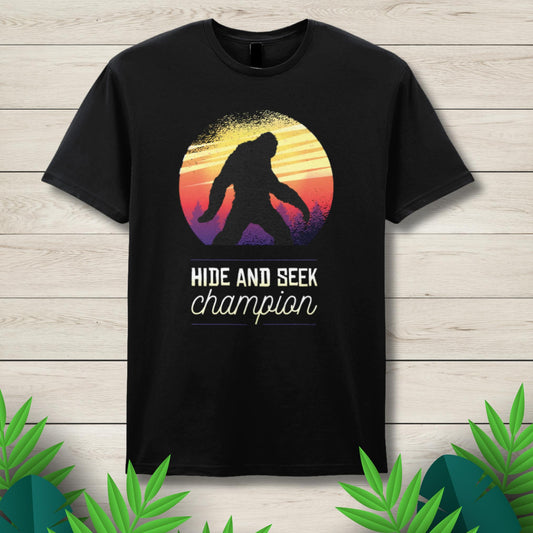 Black t-shirt with a 'Hide and Seek Champion' design featuring a silhouette of a person and a colorful sunset on a wooden background.