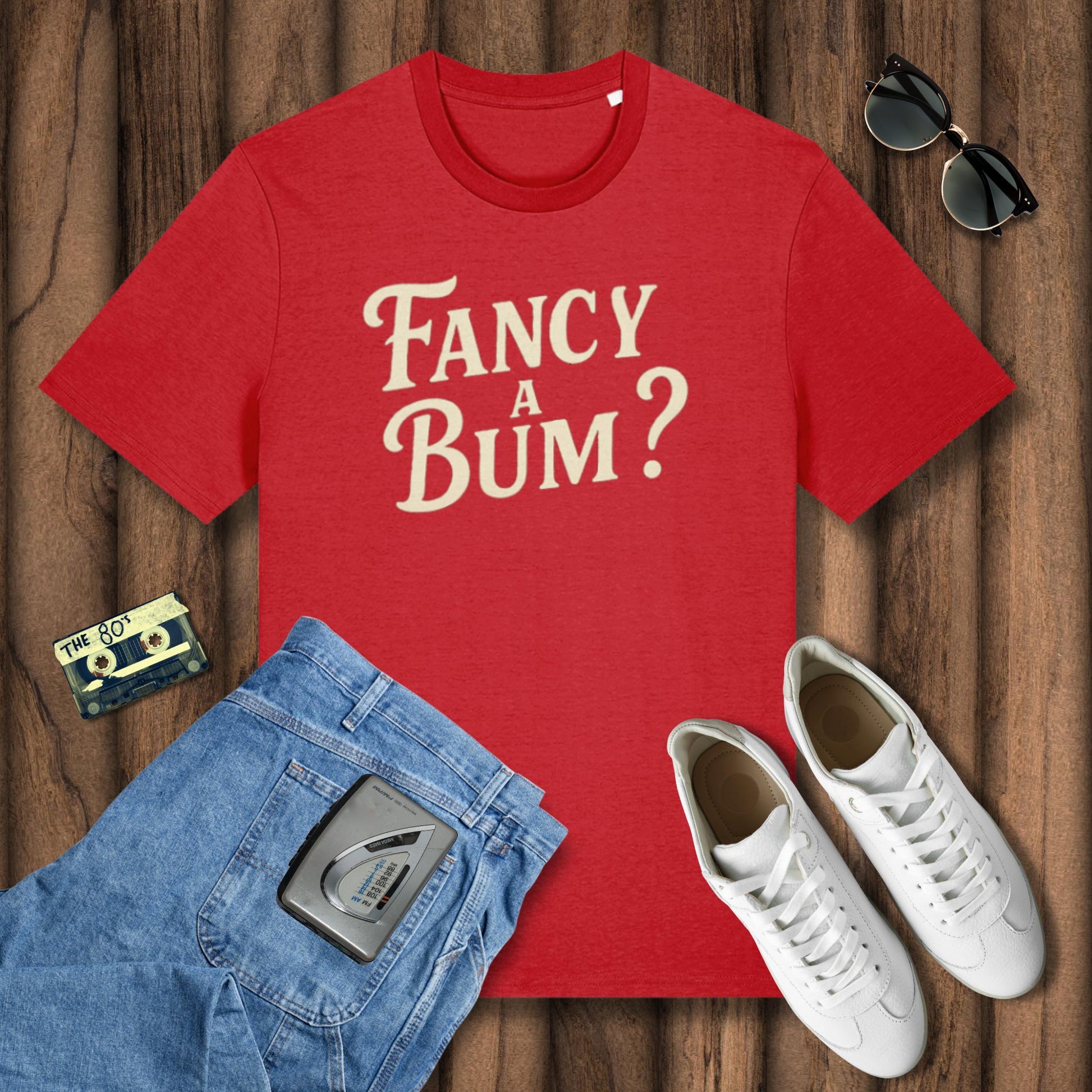 Red t-shirt with 'Fancy A Bum?' text, blue jeans, white sneakers, sunglasses, and a cassette tape on a wooden surface.