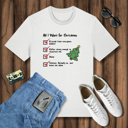White t-shirt with Christmas-themed text and graphics on a wooden surface with jeans, sneakers, and sunglasses.