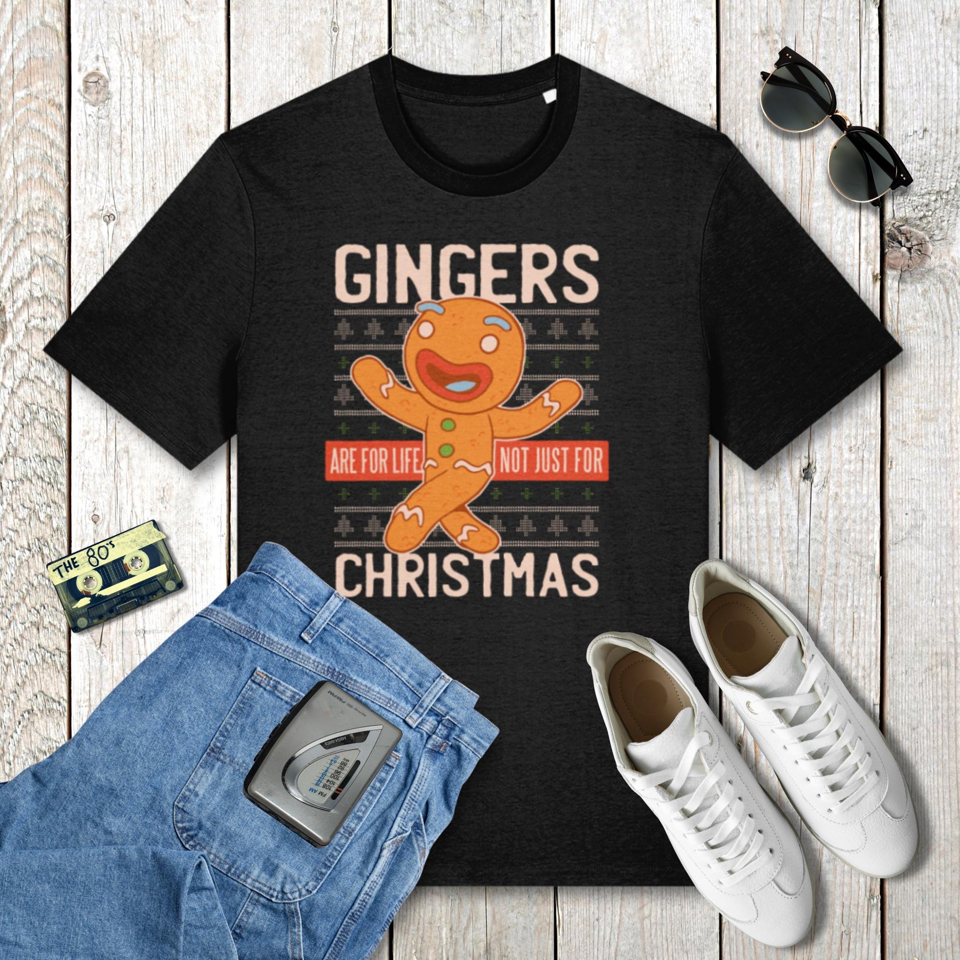 Black t-shirt with gingerbread man graphic and text on a wooden surface with jeans, sneakers, and sunglasses.