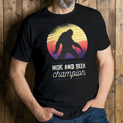man wearing a black t-shirt with a Sasquatch graphic and 'Hide and Seek Champion' text against a wooden background
