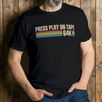 Person wearing a black t-shirt with 'Press Play on Tape 64K' text and rainbow stripes against a wooden background