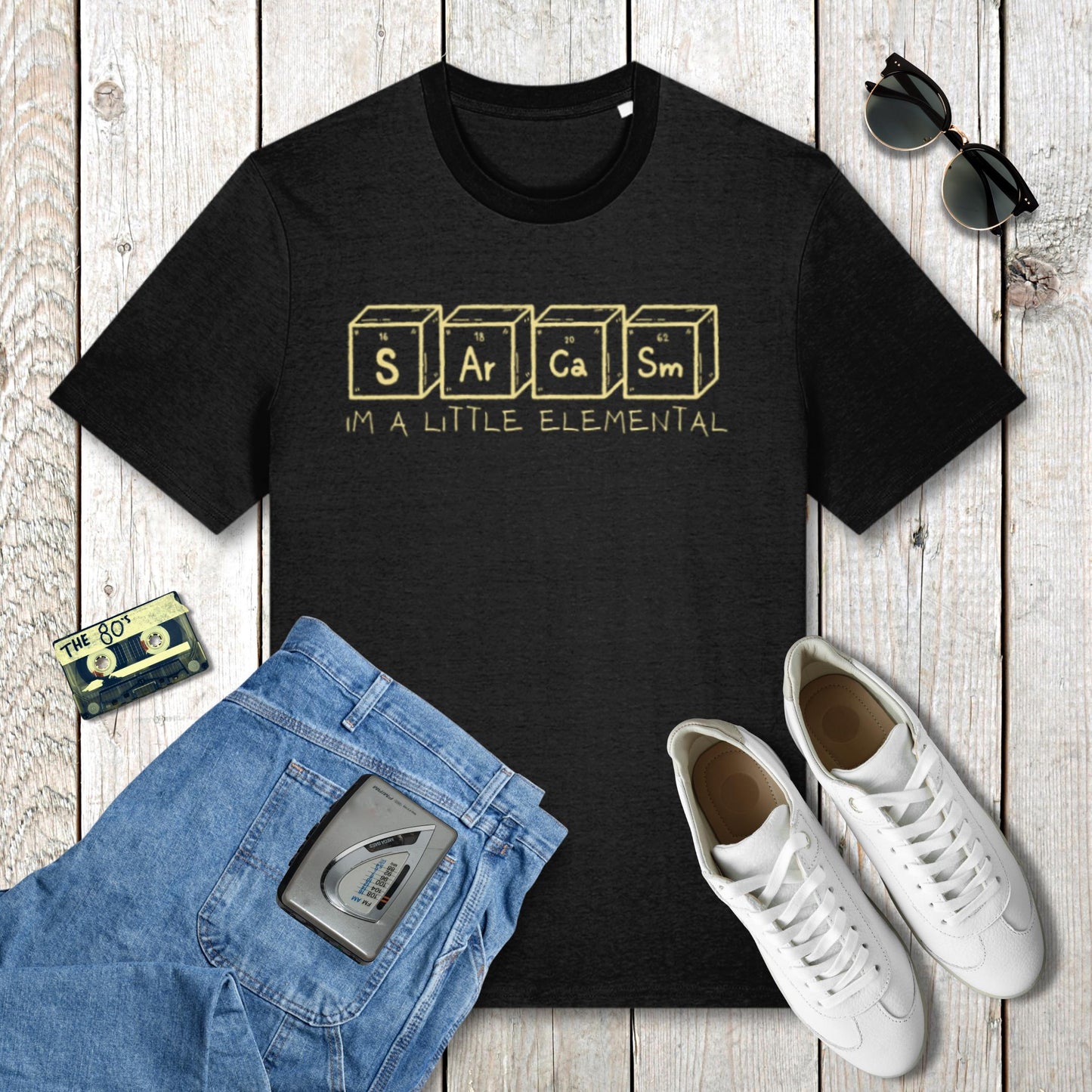 Black t-shirt with 'Sarcasm is a Little Elemental' text, blue jeans, white sneakers, and sunglasses on a wooden surface.